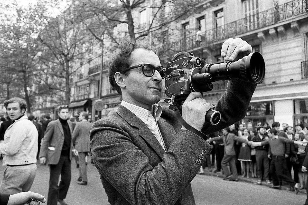 JEan-Luc Godard dans la rue en train filmer, caméra à l'épaule