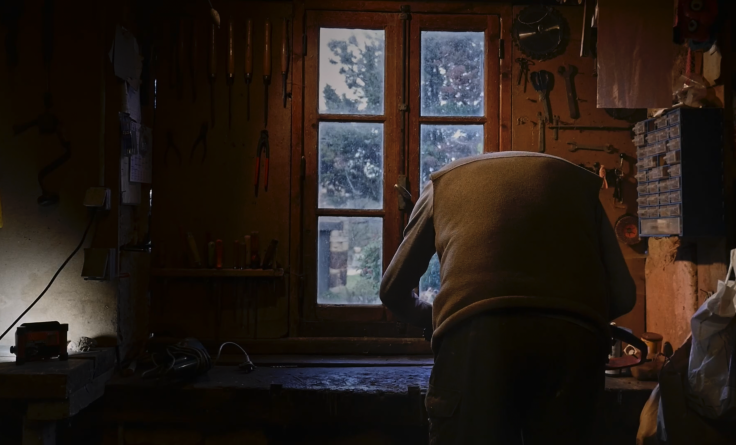 Homme de dos en train de travailler dans un petit atelier dans sa maison. Des outils sont accrochés au mur, il fait sombre.